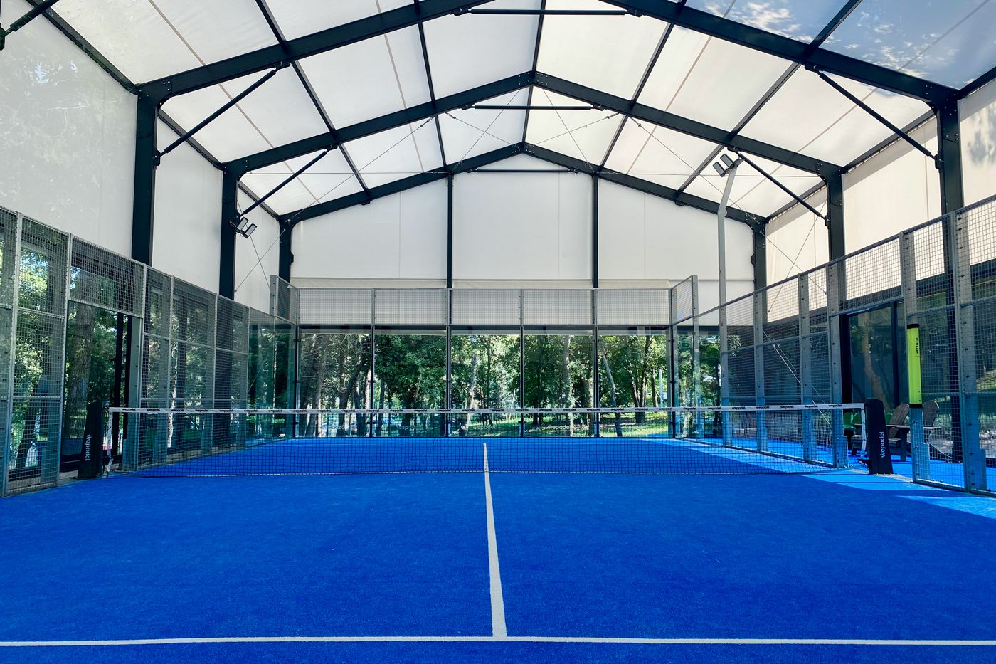 Pista de pádel cubierta vacía con suelo azul y paredes de cristal rodeada de árboles