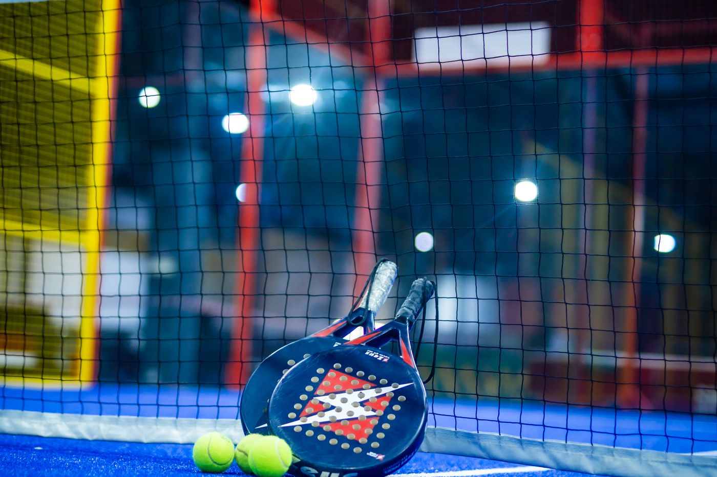 Dos palas de pádel y pelotas apoyadas en la red de una pista indoor con iluminación nocturna
