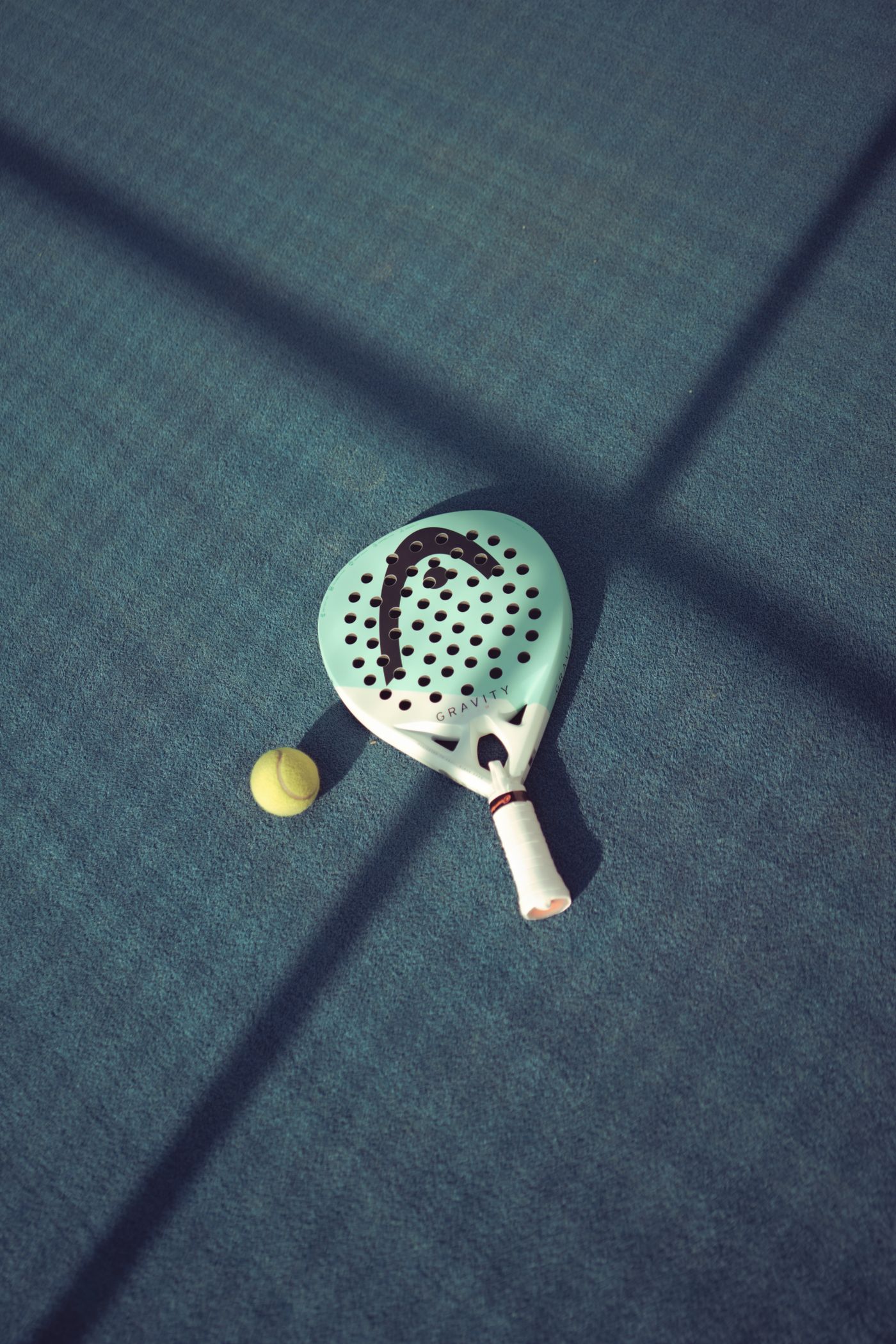 Pala de pádel apoyada sobre la pista azul junto a una pelota, luz de tarde