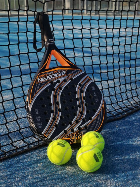 Pala de pádel y tres pelotas amarillas apoyadas en la red de una pista azul